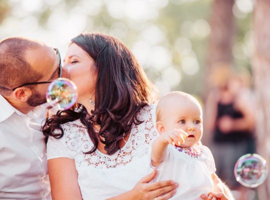 A tender family moment captured with a couple kissing and a baby holding bubbles outdoors.
