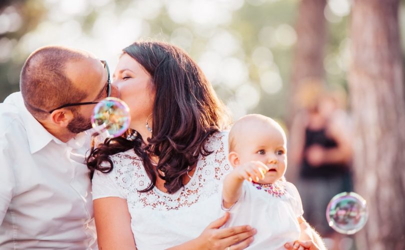 A tender family moment captured with a couple kissing and a baby holding bubbles outdoors.