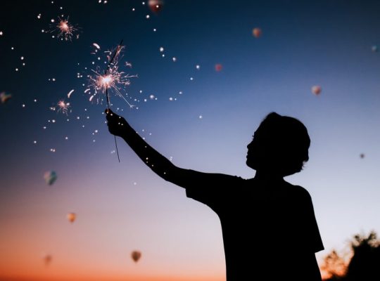 Silhouette of a person holding sparklers against a colorful twilight sky.