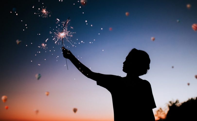 Silhouette of a person holding sparklers against a colorful twilight sky.